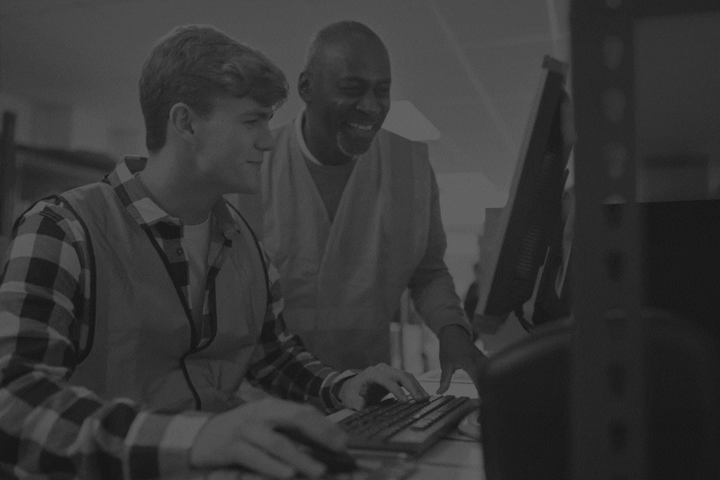 Male-Intern-With-Supervisor-Working-In-Busy-Modern-Warehouse-On-Computer