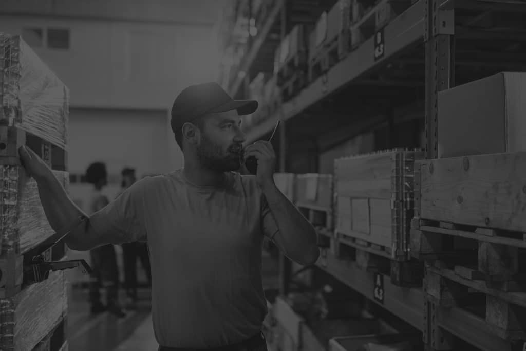 male warehouse worker communicating with someone walkie talkie while standing shelfs distribution warehouse black and white