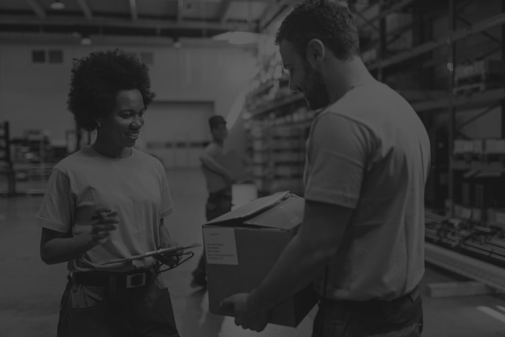 Female warehouse worker going through a checklist and talking to a coworker carrying a cardboard box, in black and white