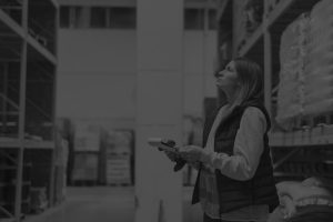 Medium shot of a woman holding a clipbooard and observing shelves in a warehouse, surrounded by pallets, captured in black and white.
