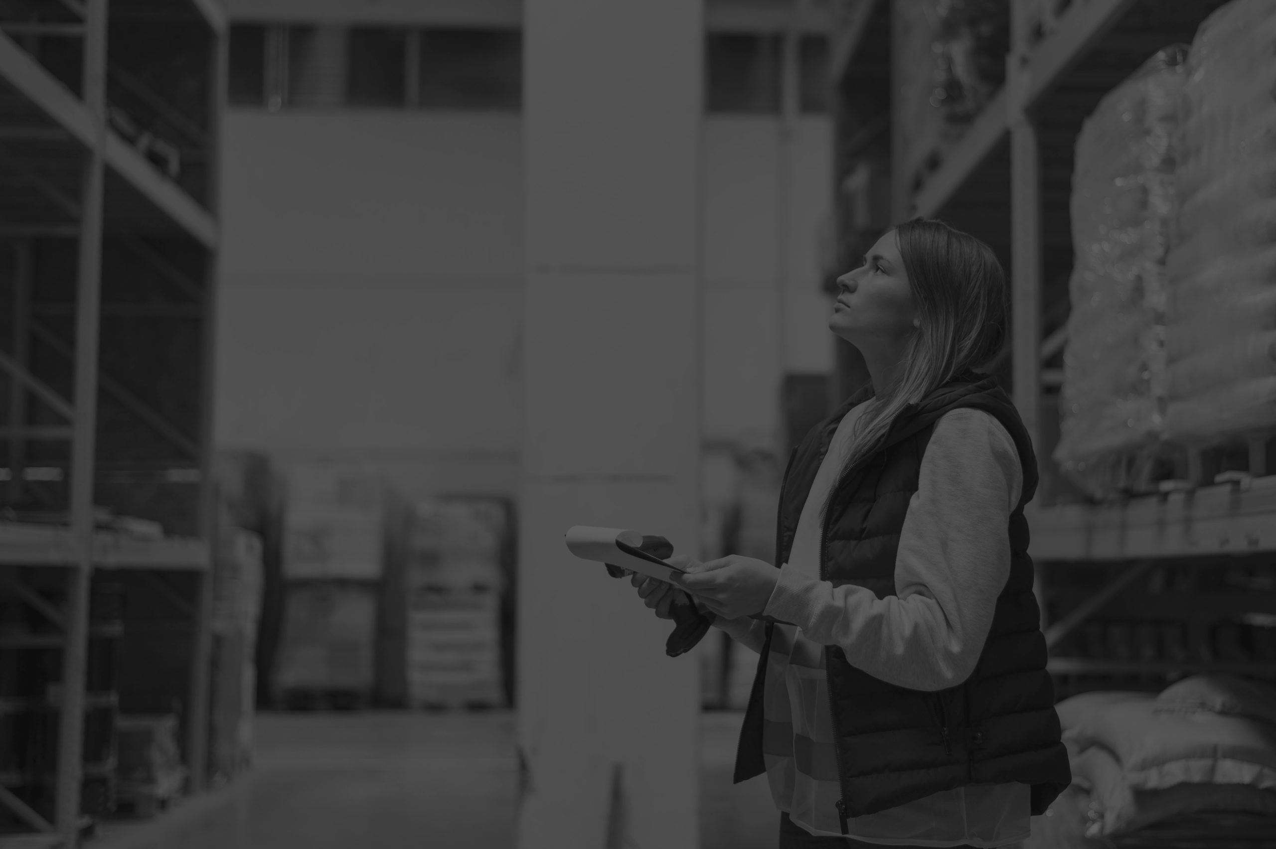 Medium shot of a woman holding a clipbooard and observing shelves in a warehouse, surrounded by pallets, captured in black and white.