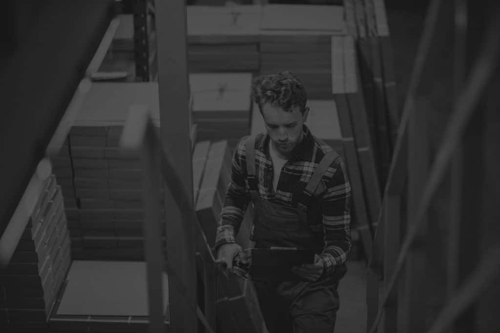 male warehouse worker climbing stairs surrounded by boxes and holding a cliboard. Black and white image.