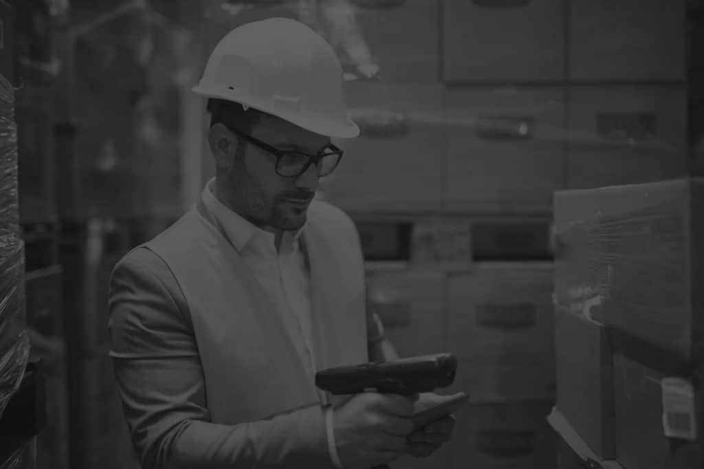 Male warehouse worker wearing hi-vis and hard hat using a barcode scanner to scan a box. Black and white image.