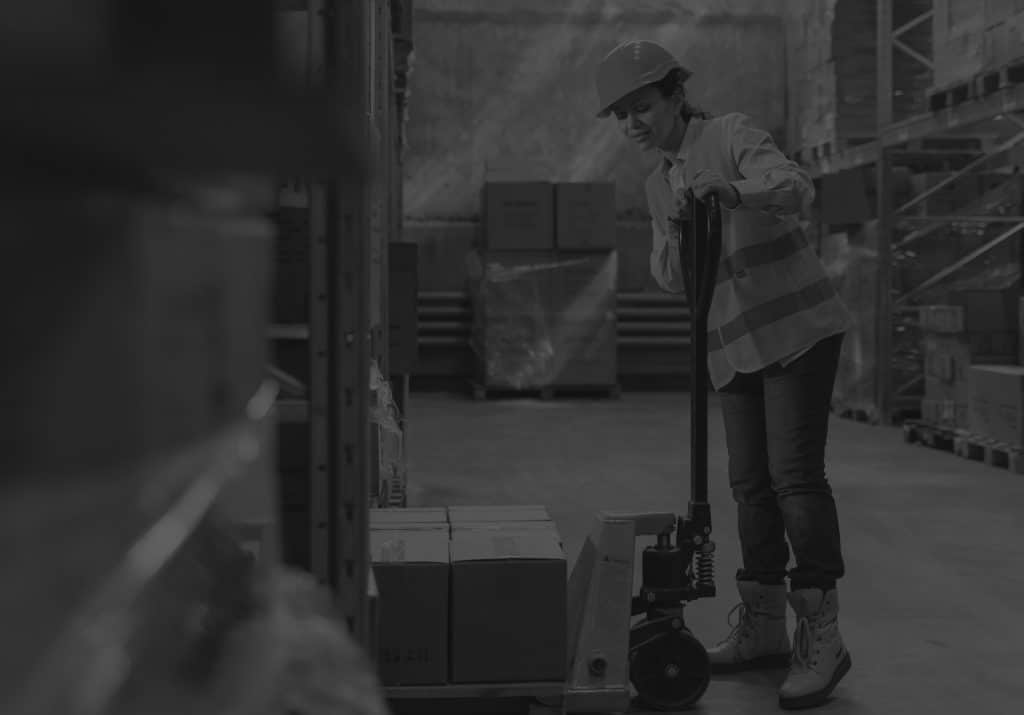 woman in warehouse wearing helmet using forklift pushing boxes. Black and white image.