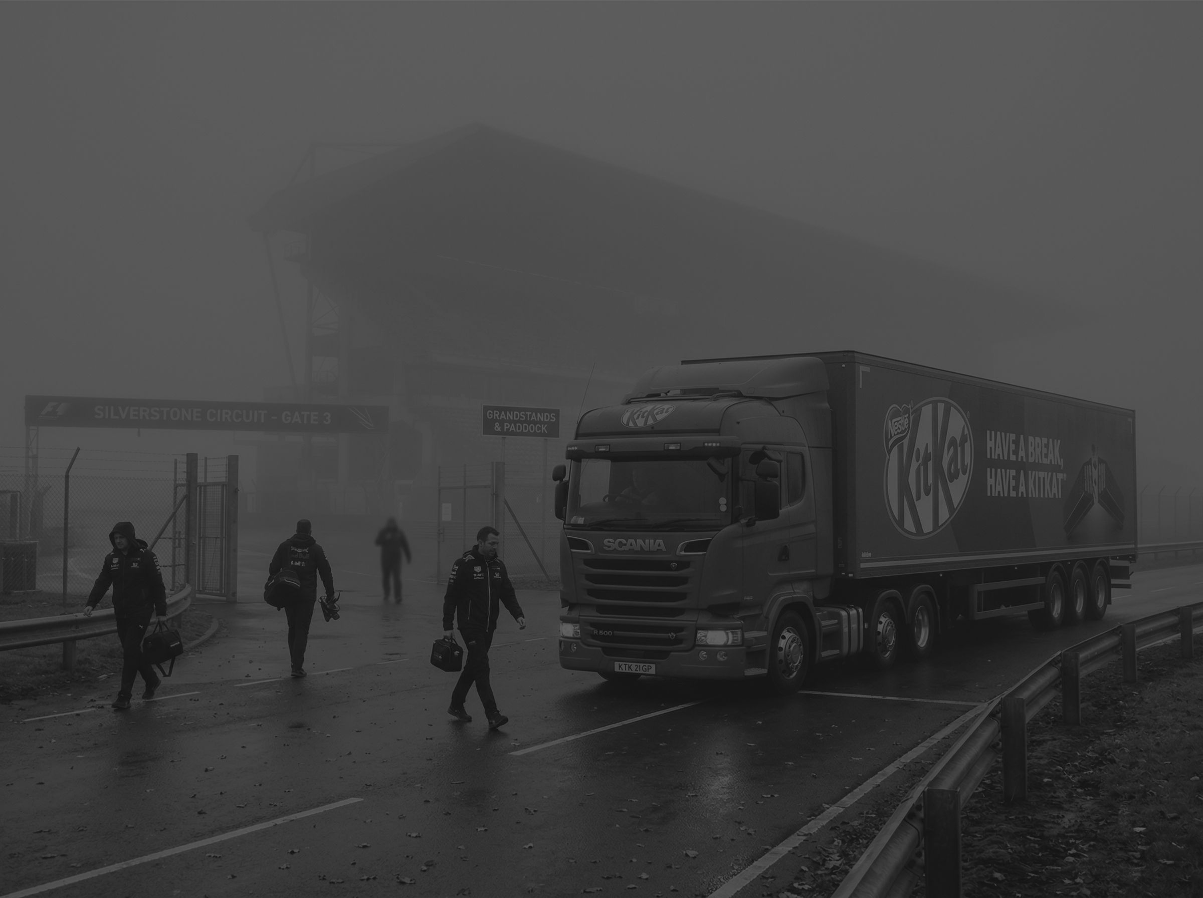 Men walking in front of parked KitKat lorry in front of formula 1 track in black and white.